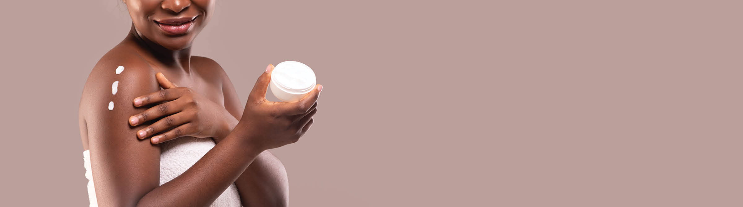 Skin Care Concept  Unrecognizable Black Lady Applying Moisturising Body Lotion After Shower, Holding Jar With Nourishing Cream, Standing Wrapped In Towel Over Gray Studio Background, Cropped Image