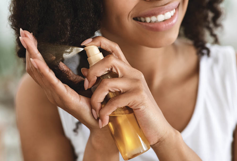 Split Ends Repair Treatment. Smiling African Woman Applying Essential Oil Spray On Her Curly Brown Hair At Home, Cropped Image, Closeup