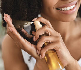Split Ends Repair Treatment. Smiling African Woman Applying Essential Oil Spray On Her Curly Brown Hair At Home, Cropped Image, Closeup