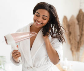 Happy Black Lady Drying Wavy Hair With Hairdryer After Shower Standing In Modern Bathroom At Home. Woman In White Bathrobe Enjoying Haircare And Styling Routine Concept