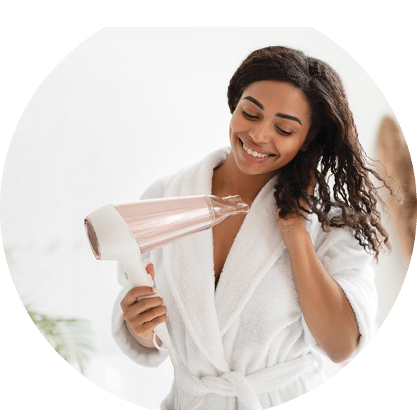 Happy Black Lady Drying Wavy Hair With Hairdryer After Shower Standing In Modern Bathroom At Home. Woman In White Bathrobe Enjoying Haircare And Styling Routine Concept