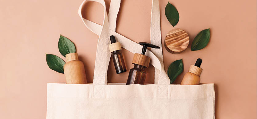 Eco-friendly cotton reusable bag with the different tubes and bottles from the natural wood and brown glass.Fresh natural leafs around.Concept of the organic,zero waste cosmetics.
