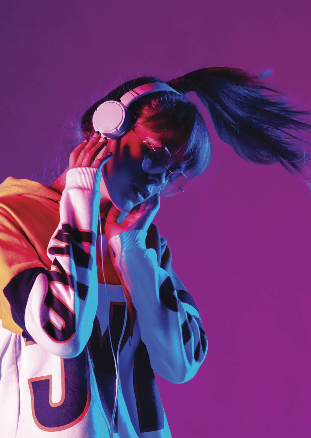Stylish fashion teenager model wearing hoodie and headphones listening dj music dancing in purple neon lights. Young teen girl enjoy cool music 90s party mix in violet studio background. Copy space.