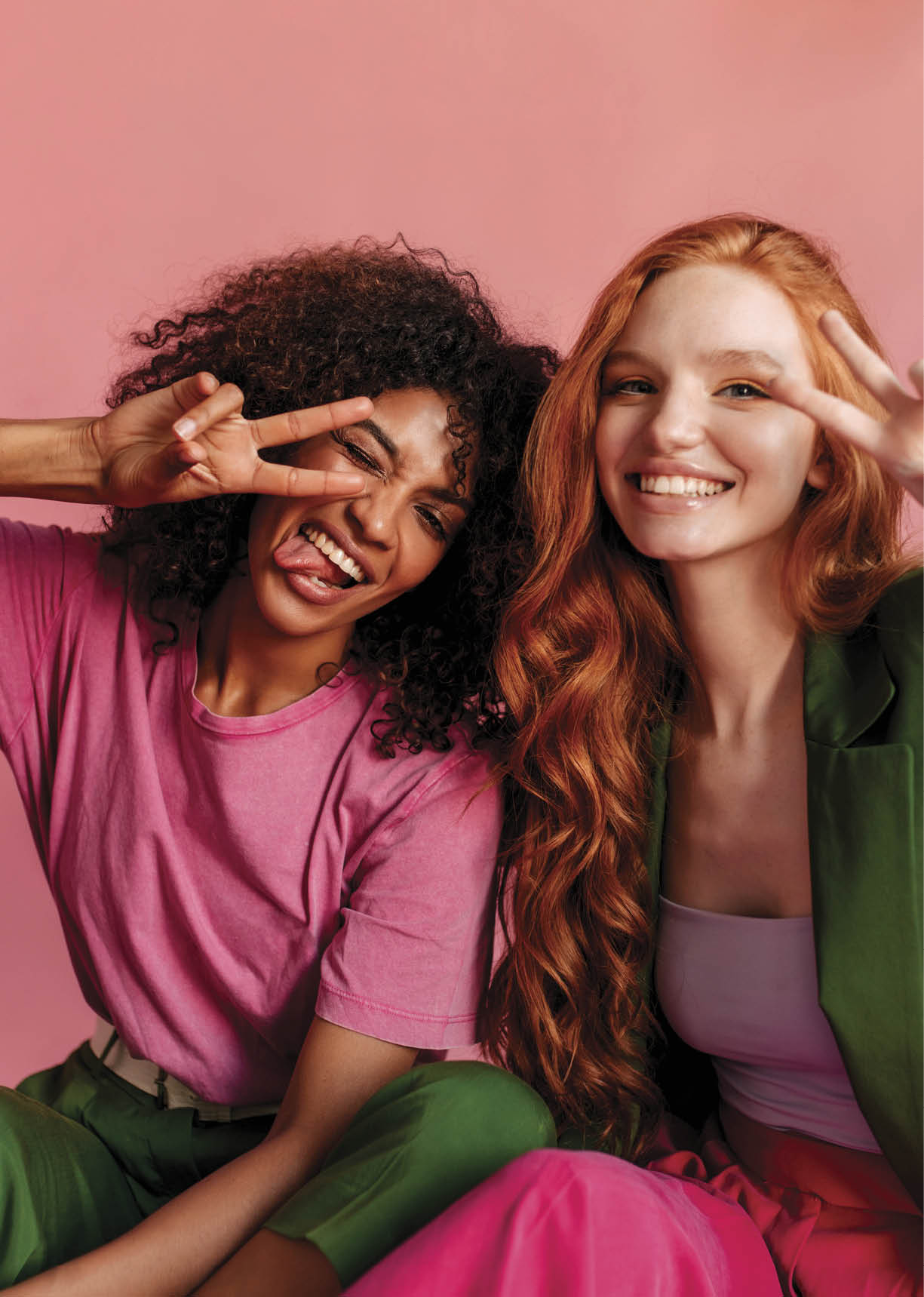 Cheerful young international women demonstrate peace gesture against pink studio background. African brunette in T-shirt, her tongue sticking out next to european model with long wavy red hair.