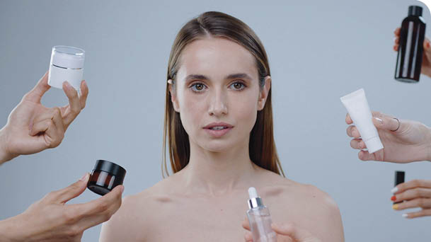 Close up of beautiful young woman look at camera on a grey background around a lot hands hold cosmetics appear and disappear attractive care clean complexion eyes face girl health makeup slow motion
