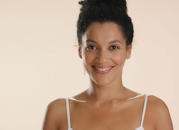 Beauty portrait of young positive good-looking African American woman with dark curly hair in bun in white top smiling wide for the camera on beige background | Unwanted arm hair removal concept