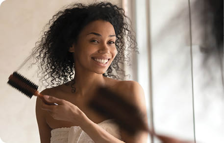 Happy african ethnicity millennial wrapped in towel after morning shower or bath woman standing in bathroom, looking in mirror, brushing, styling, take care of curly hair. Natural beauty care routine.